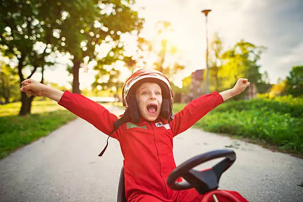 Little boy aged 6 has won a a pedal go-kart race (or kart, soapbox car, cyclekart race). Little boy is happily rising his arms in winning gesture. Road and sunset visible in the background.