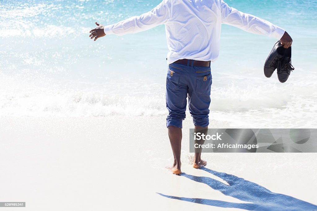 African male walking towards the sea water fully clothed. African male on the sea shore walking towards the sea water with his arms open wide, fully clothed and his shoes in his right hand. Cape Town, Western Cape, South Africa. 20-24 Years Stock Photo