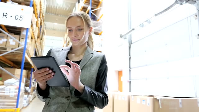 Hd slow motion video of young female manager of large distribution warehouse using digital tablet and standing in aisle of futuristic factory. Beautiful supervisor holding digital tablet and checking orders. On-line goods orders worldwide. Blond concentrated worker wearing elegant suit, looking successful and confident. Large distribution warehouse in background with forklift, racks full of packages, boxes, pallets, crates ready to be delivered. Logistics, freight, shipping, receiving.