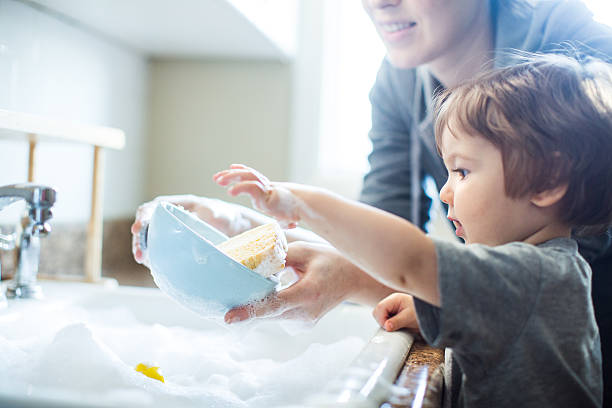 Baby Dish Washing A cute toddler aged boy wearing a blue diaper and tshirt helps out with with washing the dishes, standing on a chair to be able to reach the sink full of soapy bubbles and dishes.  His mother smiles behind him as she supervises his sensory perception experience. Bright sunlight comes in through the window behind him, lighting the sparse modern kitchen. leanincollection stock pictures, royalty-free photos & images