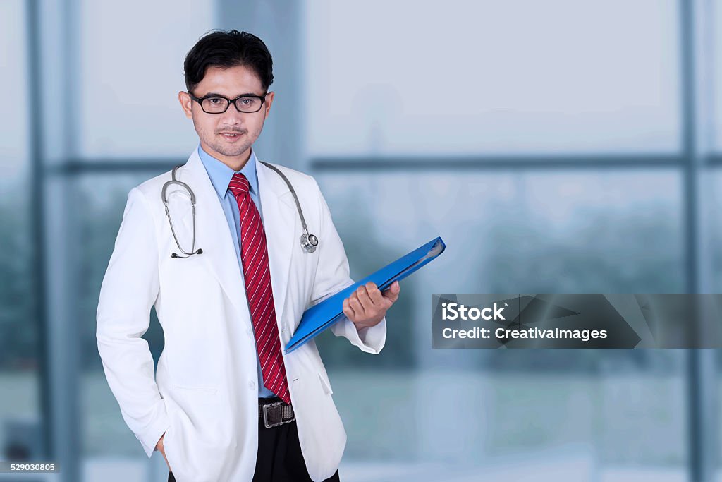 Handsome doctor standing near the window Young asian doctor wearing coat and smiling at the camera while holding a document near the window Adult Stock Photo