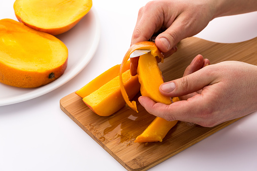Skin of a mango wedge is being separated from the juicy flesh of this tropical fruit. One hand holding the fruit chip and the other operating a kitchen knife. Wooden board dabbled with mango juice.