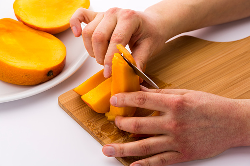 Blade of a short kitchen knife is separating the fruit flesh from the skin of a mango wedge. A left hand is holding the mango chip in place, while a right hand is driving the knife. White background.