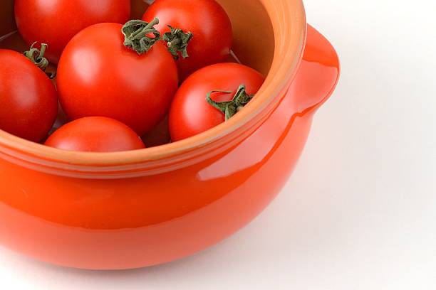 A rustic ceramic casserole overflows with glossy fresh green cherry tomatoes on the sunlit linen woven carpet, lid casually placed nearby, beneath a soft white linen cloth on the wall.