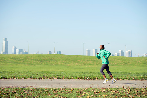 Runner woman jogging on asphalt road in green city park. Female jogger in outdoor run jacket training outside. Blue skyline is on background.