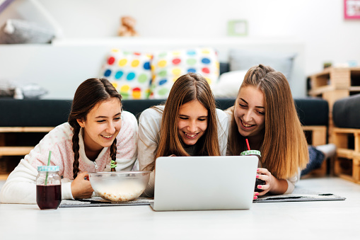 Teenage Girls Having fun together at home. They are using laptop on slumber party. Wearing casual clothes and enjoying. Lying on the floor on front and drinking juice from the jars. There is a bowl with popcorn in front of them.