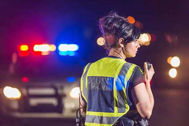 Rear view of a female police officer standing in the street at night, talking into her radio. Her patrol car is in the background with the emergency lights illuminated. She is wearing a yellow safety vest