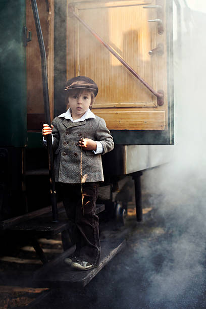 Boy, dressed in vintage coat and hat, standing on the stairs of steam train, holding pocket watch, steam behind him