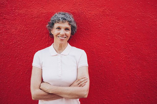 Portrait of happy mature woman with her arms crossed standing against red background. She is leaning to a red wall with copy space.