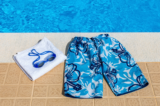 Blue swimming trunks goggles and bath towel at pool. Water in blue swimming pool with swimming stuff  on the edge of the terrace. It's summer season. On this warm sunny day in summer season it's nice to cool down and swim.