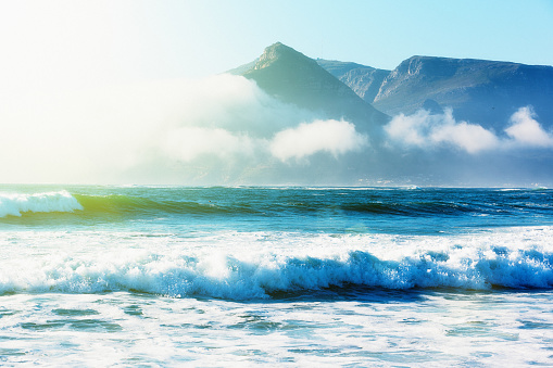 The sun shines on gentle surf; in the background, mountains are surrounded by fluffy clouds. Shot in South Africa's Western Cape.