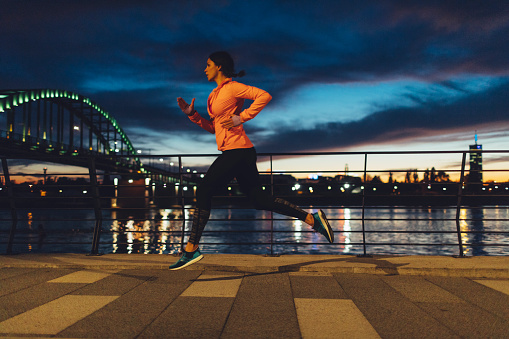 Young Woman Jogging Near River at night. She is wearing modern sporty clothing. With bridge and cityscape in background.