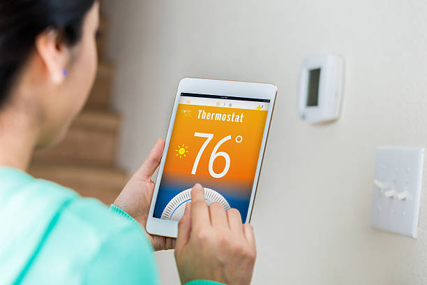 Woman uses digital tablet to control home's temperature Female homeowner uses technology on digital tabelt to control her home's temperature remotely. She is standing near the thermostat's control panel. View is over the woman's shoulder. Focus is on the digital tablet. smart thermostat photos stock pictures, royalty-free photos & images