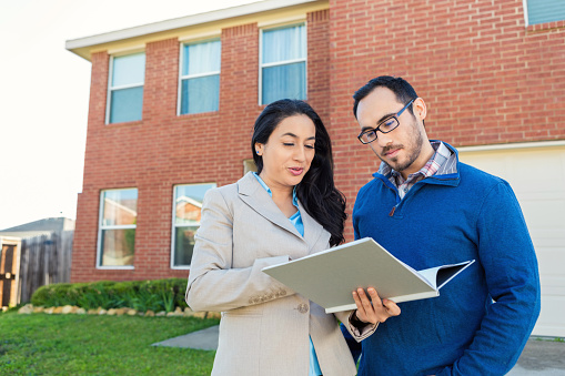 Mid adult Hispanic real estate agent reviews paperwork with potential client or new homeowner in front of red brick two-story home with front entry garage. They are both wearing business casual attire.