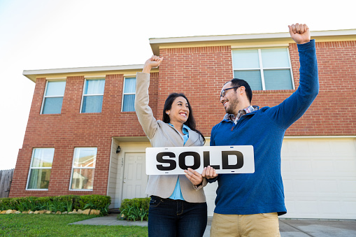 Mid adult Hispanic new homeowners or husband/wife real estate team celebrate new home purchase. They are cheering with their arms raised and are holding a 'sold' sign. Red brick two-story home  with front entry garage is behind them. They are dressed in business causal clothing.