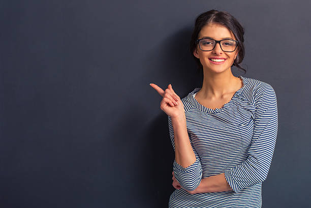 Attractive young woman Attractive girl in casual clothes and eyeglasses is pointing away, looking at camera and smiling, standing against dark background beckoning photos stock pictures, royalty-free photos & images