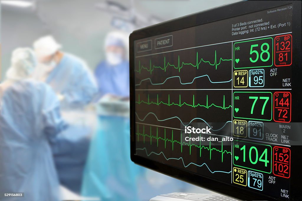 Intensive care unit monitor and surgery Intensive care unit (ICU) LCD monitor with an ongoing surgery in background Electrocardiography Stock Photo