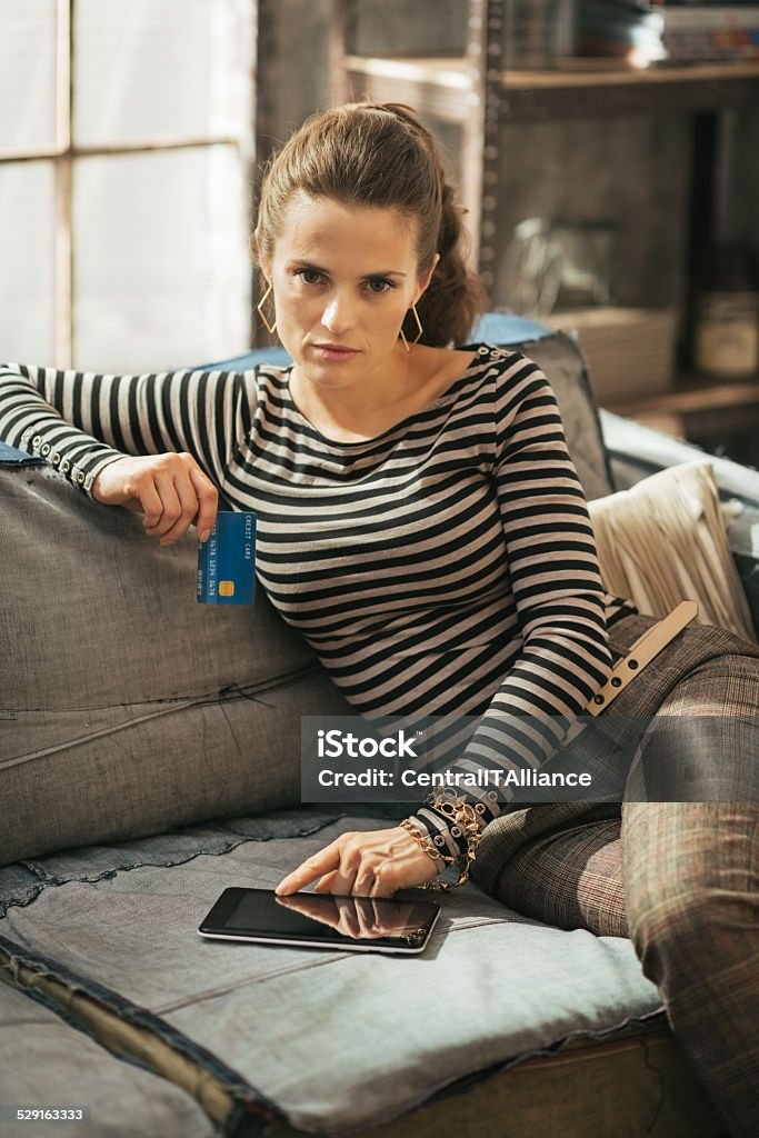 Woman with credit card and tablet pc in loft apartment Thoughtful young woman with credit card and tablet pc sitting on divan in loft apartment Adult Stock Photo