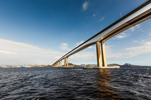 A DSLR photo of Rio Niteroi Bridge from inside Guanabara Bay in Rio de Janeiro, Brazil. The bridge is seen from a low angle view in a diagonal way from the up right corner to the midle left of the frame. It is a sunny afternoon with blue sky and scattered white clouds.