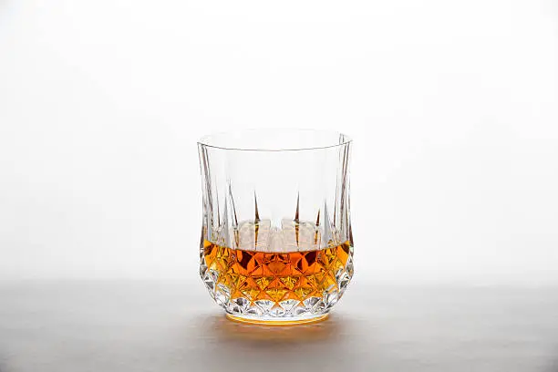 A single diamond-cut tumbler containing whiskey, bourbon, brandy, cognac, scotch, or other amber liquid is sitting on a cool grey background with copy space at the top of the frame.  The image could apply to numerous concepts including – bar and restaurant themes, alcohol themes, celebration, relaxation, drinking, and many more.