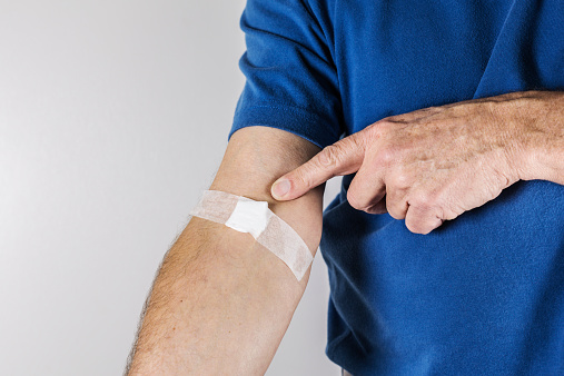 A senior man health labratory patient in a blue polo shirt has just given blood for testing. He is pointing with his finger to the white bandage tape and sterilized gauze on his arm near the inside of his elbow.