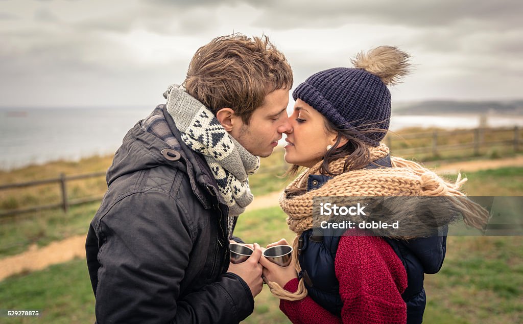 Young couple kissing and holding cups of hot drink outdoors Portrait of young happy couple kissing and holding cups of hot beverage in a cold day with sea and dark cloudy sky on the background Adult Stock Photo