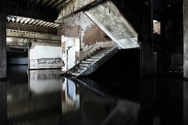 This is a horizontal, monochromatic photograph of an abandoned building interior. Water floods the bottom. A staircase leads up in the center. Photographed with a Nikon D800.