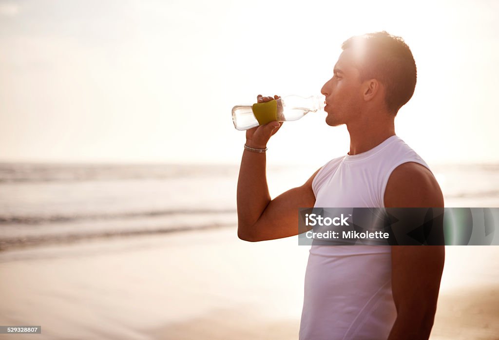 Post-run refreshment at sunset Shot of a young man drinking from his water bottle while out for a run on the beach Activity Stock Photo