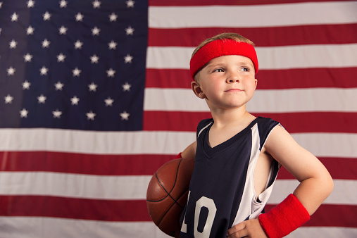 Portrait of a young boy dressed in his basketball uniform standing with his basketball in front of a giant American flag. This boy is ready to show his skills on the court.