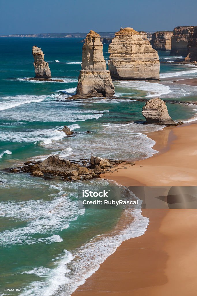 Vertical scenic view of The Twelve Apostles in daylight The Twelve Apostles, Great Ocean Road, Victoria, Australia. Scenic, vertical view overlooking the coastline from the cliff top. Taken in the harsh, bright, daytime light. There are four sea stacks in view, with surf waves rolling in with lots of froth. The sand is golden-yellow and the sea bright green.  Australia Stock Photo