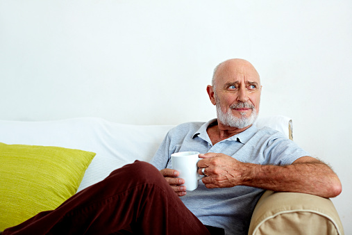 Thoughtful senior man holding coffee mug while relaxing on sofa at home