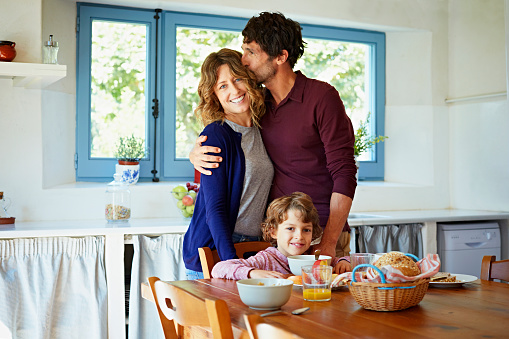 Loving man kissing woman while standing behind son having breakfast at table in kitchen