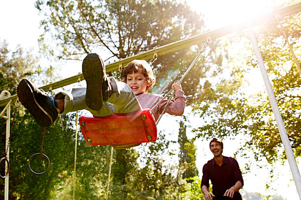 father pushing son on swing in park - swinging imagens e fotografias de stock