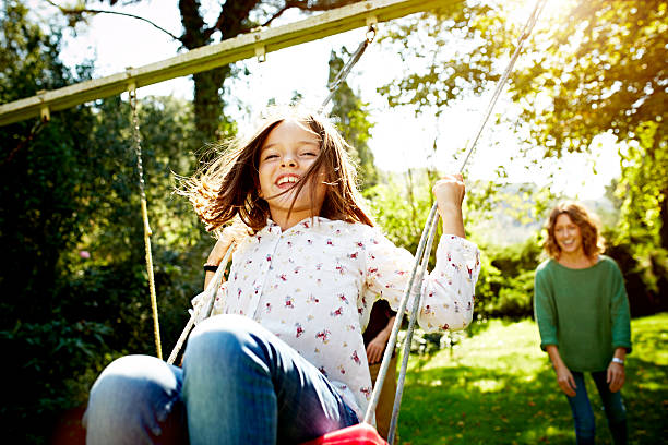 Mother pushing daughter on swing in park Cheerful mother pushing daughter on swing in sunny park swing play equipment stock pictures, royalty-free photos & images