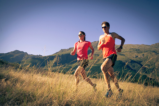 A couple run together up a grassy hill in the mountains. The couple are wearing brightly coloured running tops.