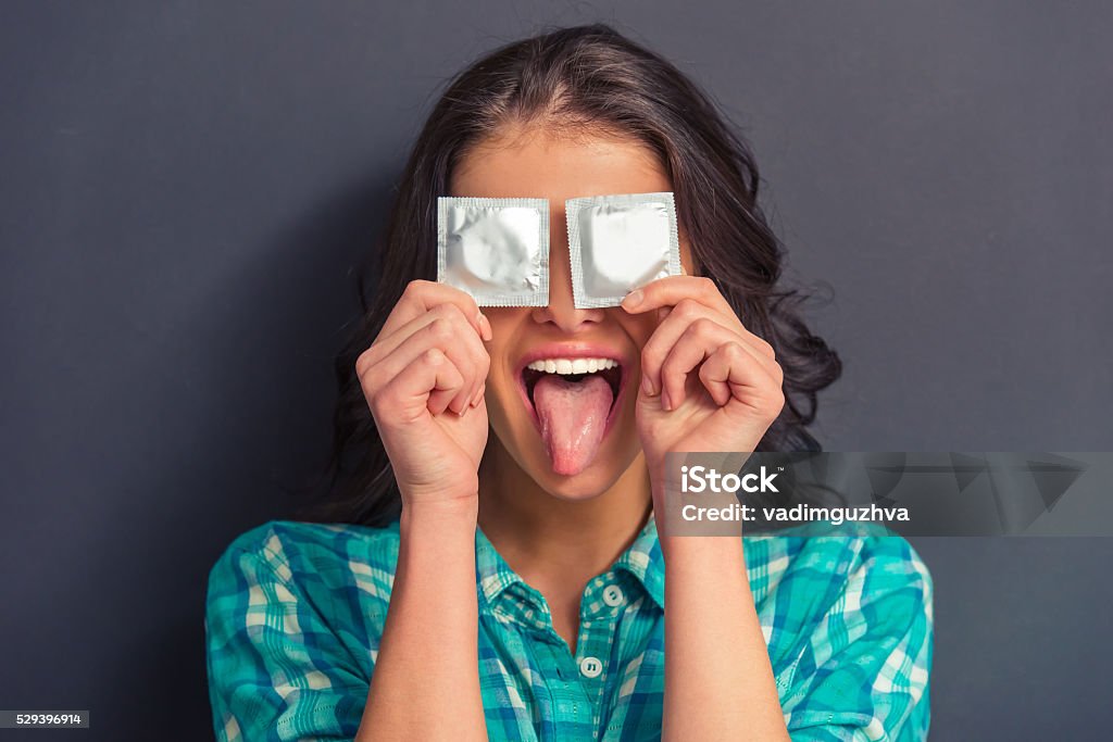 Attractive young woman with condom Portrait of attractive girl covering eyes with condoms and showing her tongue, against dark background Condom Stock Photo