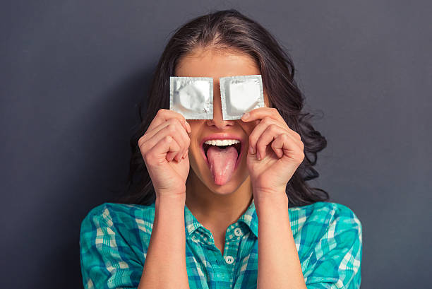Attractive young woman with condom Portrait of attractive girl covering eyes with condoms and showing her tongue, against dark background condom photos stock pictures, royalty-free photos & images