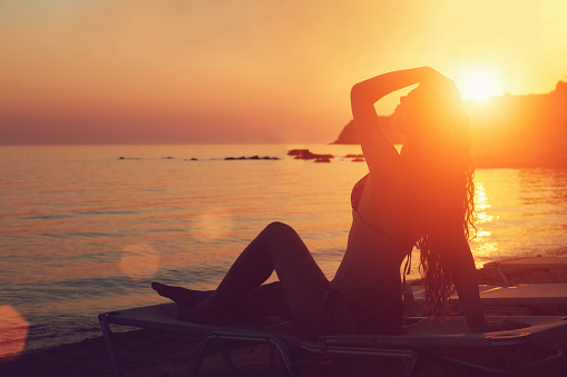 Silhouette of an attractive young woman resting on a sunbed, enjoying a beautiful summer day at the beach.