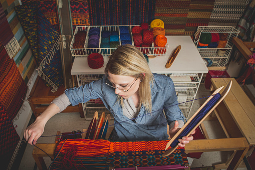 A young creative girl working on a loom surrounded by beautiful woven rugs. The young woman is in her 20's and is confidently working on the loom (stock image).