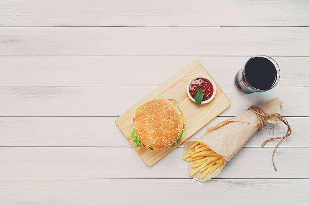 Burger, chips and cola at white wood Burger, Chips and cola. Hamburger and french fries wrapped in brown wrapping paper. Fast food take away at white shabby chic wood. Hamburger with tomato sauce. Top view, flat lay with copyspace white shabby chic desk stock pictures, royalty-free photos & images