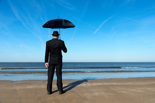 Englishman with bowler hat and umbrella stands  on the beach overlooking the sea and towards Europe.