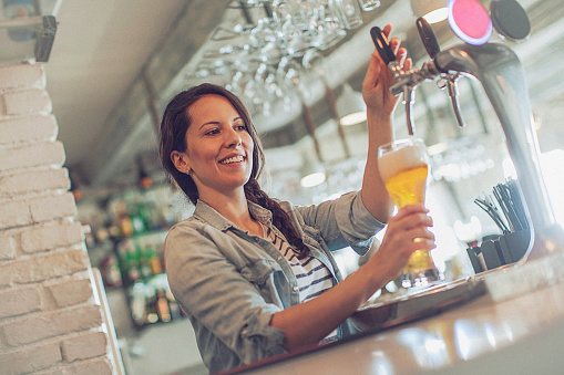 Casual young cheerful female bartender pouring beer from beer tap. Focus on the beer glass.