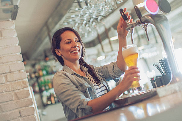Casual young cheerful female bartender pouring beer from facet stock photo