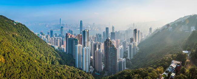 Early morning sunlight flaring over the green forested hills of Hong Kong island high above the towering skyscrapers, high rise apartment buildings and crowded futuristic cityscape that surrounds Victoria Harbour, China. ProPhoto RGB profile for maximum color fidelity and gamut.