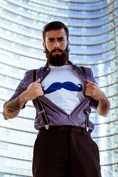 Young man (hispter style) with big earring and full beard, showing mustaches on white t-shirt with business office on background (Hipster Superhero) - (Italy - Milan - Gae Aulenti Square)
