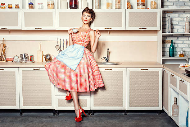 Woman in the kitchen. Woman in an apron in the kitchen drinking from a cup and waving. pics for classic pin up poses stock pictures, royalty-free photos & images