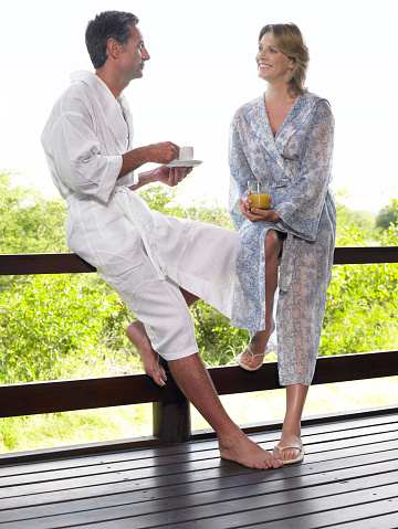 Man and woman in bathrobes leaning on terrace railing with tea