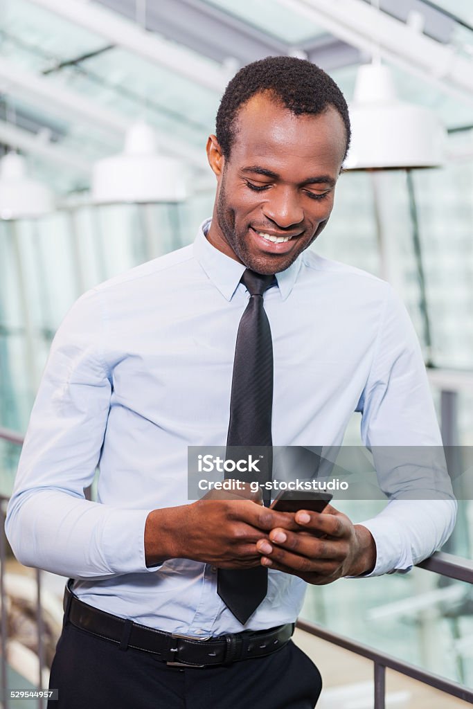 Typing business message. Cheerful young African man in shirt and tie holding mobile phone and looking at it with smile while standing indoors Adult Stock Photo