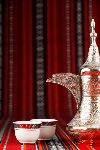 Ornate Arabian tea cups and a dallah are placed on traditional red fabric from the Gulf region.