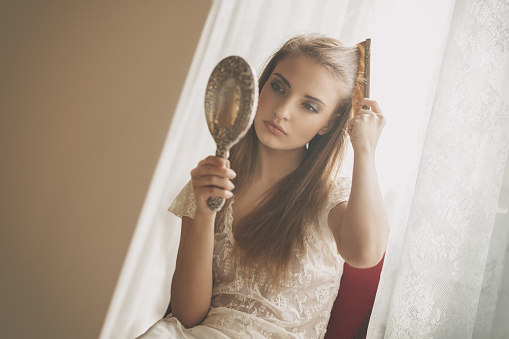 young beauty combing by the window, holding silver hand mirror and comb